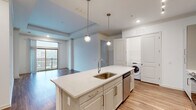 Located at 2925 Buford Dr in Buford, Georgia, this single-floor unit offers a thoughtfully designed open-concept layout with contemporary finishes throughout. The kitchen serves as a central focal point, anchored by a large island with a quartz countertop, undermount sink, and pendant lighting above. White shaker-style cabinetry lines the walls alongside a hexagonal tile backsplash, and stainless steel appliances—including a range, over-the-range microwave, and French door refrigerator—complete the space. An in-unit washer is conveniently positioned nearby. Light wood-tone flooring flows through the kitchen and living areas, while a large sliding glass door opens to a private balcony, bringing in natural light and outdoor views. The bedrooms are carpeted and feature ceiling fans and generously sized windows that overlook the surrounding area. The bathrooms are finished with clean, modern details: one includes a glass-enclosed walk-in shower with tile surround, while another features a soaking tub set within full-height stone-look tile walls. The overall ambiance is bright, neutral, and move-in ready.