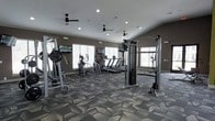 This fitness center is designed to provide a comprehensive workout experience in a spacious, well-appointed environment. The room is outfitted with an array of modern exercise equipment, including multiple treadmills, elliptical machines, and cable strength training stations, offering members a full range of cardio and resistance training options. Patterned carpet tiles in a geometric, multi-toned gray design cover the floor, adding both visual interest and comfort underfoot. Multiple ceiling fans are evenly distributed throughout the space, ensuring consistent airflow and a comfortable workout environment. Recessed lighting in the white ceiling provides bright, even illumination throughout. Large windows line the walls, allowing ample natural light to fill the room while offering views to the outside. The walls are finished in a warm taupe tone, complementing the contemporary aesthetic of the space. Wall-mounted flat-screen televisions are positioned at strategic viewing angles, allowing members to stay entertained during cardio sessions. The overall ambiance is clean, modern, and functional, making this fitness center a well-rounded facility suited for a variety of fitness routines.