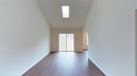 This two-floor space is defined by its striking architectural design, most notably the vaulted ceilings that soar overhead in the main living area, creating an expansive sense of volume and openness. A centrally positioned skylight floods the space with natural light from above, complementing the large sliding glass door along the rear wall that provides additional brightness and outdoor access. The living area is finished with warm-toned hardwood-style flooring that runs throughout, contrasting cleanly against the neutral, light-colored walls. The clean lines of the white baseboards and the smooth, textured ceiling contribute to the space's understated, modern aesthetic. An adjacent room is partially visible through an open doorway to the right, benefiting from its own window that introduces further natural light. The overall ambiance is clean, airy, and versatile, with the high ceilings and abundant light sources making the main living area feel particularly spacious. The two-floor layout offers additional functional living space beyond what is visible, providing room for a variety of living arrangements throughout the home.