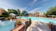 This single-story clubhouse facility is set against the backdrop of a stately, multi-story brick building with a distinctive green roof and large white-framed windows. The exterior is highlighted by a generously sized outdoor swimming pool with clearly marked lap lanes, surrounded by a spacious brick and stone pool deck furnished with lounge chairs and shade umbrellas. Mature trees frame the property, adding a lush, landscaped ambiance to the outdoor area.

The interior entry features terracotta brick-patterned tile flooring and crisp white walls accented with dark wainscoting and bold black trim, lending the space a clean, classic aesthetic. Large glass windows near the entry allow natural light to filter in and provide views of the surrounding grounds, including a glimpse of an interior staircase. A conveniently located snack and beverage vending machine offers easy access to refreshments. Separate men's and women's restrooms are situated just off the main entry corridor, each clearly marked with signage and framed by dark-trimmed white panel doors that are consistent with the overall design language. The overall ambiance is polished and functional, balancing traditional architectural details with practical amenities.