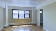 This single-floor space showcases a clean, functional design with notable classic details throughout. The main living area is defined by warm parquet wood flooring arranged in a traditional basket-weave pattern, lending a sense of timeless character to the space. Large windows fitted with horizontal blinds allow generous natural light to filter in, brightening the room and highlighting the flooring's natural tones. The walls are finished in a neutral off-white and warm yellow palette, creating an open and inviting atmosphere. A baseboard heating system runs along the perimeter of the room, offering an efficient and unobtrusive heating solution. The layout flows naturally into an adjoining room, visible through a wide doorway framed in crisp white trim — that secondary space also features a window, maintaining consistent natural light throughout. The overall design is straightforward and practical, with clean lines and well-maintained finishes. The combination of parquet flooring, ample windows, and neutral tones creates a versatile environment well-suited for a variety of uses.