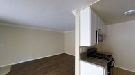 This single-floor residence offers a practical and well-appointed layout that seamlessly connects the kitchen and living areas. The open design promotes a sense of spaciousness, with warm, neutral-toned walls complemented by crisp white crown molding that adds a clean, classic touch throughout. Rich, dark hardwood-style flooring anchors the living space, providing a striking contrast against the light-colored walls.

The kitchen is equipped with white cabinetry that extends to the ceiling, maximizing storage capacity. Granite countertops in earthy tones provide a durable and stylish workspace, paired with stainless steel appliances including an over-the-range microwave and a gas range. A structural column with a decorative crown detail tastefully defines the boundary between the kitchen and the adjacent living area without fully closing off the space.

The living area is generously proportioned, offering ample room for flexible furniture arrangements. A textured ceiling adds subtle character overhead, while the consistent flooring and neutral wall palette throughout create a cohesive and inviting ambiance. The overall design balances functional efficiency with understated elegance, making this a comfortable and move-in ready space.