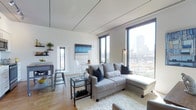 This single-floor space showcases a thoughtfully designed open-plan layout that seamlessly integrates the living and dining areas. The interior is defined by its bright, white walls and smooth ceilings fitted with subtle architectural track lighting, which complement the warm hardwood flooring beneath. Floor-to-ceiling windows dominate one wall of the living area, flooding the space with natural light while framing impressive panoramic city skyline views and a private balcony beyond.

The living area is generously proportioned, centered around a plush gray sectional sofa with chaise, paired with a glass-and-metal coffee table that adds an industrial yet refined touch. A soft area rug anchors the seating arrangement, enhancing the sense of comfort. Decorative wall art in blue and neutral tones adds a curated aesthetic.

Adjacent to the living space, a casual dining area features a round table with ghost chairs, maintaining the open and airy feel. A compact shelving unit with greenery introduces natural elements. The overall ambiance is clean, contemporary, and urban, making effective use of natural light and city views to create an elevated, modern living environment.
