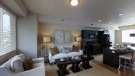 This three-floor space presents an open-concept layout that seamlessly connects the living area and kitchen, creating a functional and visually unified environment. The living area is anchored by a plush, light-toned sofa adorned with decorative pillows in neutral and earth tones, complemented by a pair of matching armchairs. A set of dark-finished, geometric coffee tables sits atop a textured area rug, adding visual depth to the room. Ambient lighting is achieved through a combination of recessed ceiling lights, a central flush-mount fixture, and tall floor lamps with warm-toned shades, contributing to the inviting atmosphere. A large abstract artwork serves as a focal point on the main wall, enhancing the contemporary aesthetic. The kitchen, visible from the living space, features dark cabinetry paired with black appliances, including a range and overhead microwave, providing a bold contrast to the lighter tones of the living area. The overall color palette throughout is composed of warm neutrals, grays, and deep browns, producing a cohesive and sophisticated ambiance across the space.