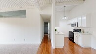 Located along Pacific Coast Highway in Torrance, California, this single-floor residence is set within a multi-story building surrounded by lush palm trees and mature landscaping. The unit's most distinctive architectural feature is its white-painted exposed beam and plank ceilings, which lend a mid-century character and a sense of openness throughout the space. Hardwood flooring runs through the main living and kitchen areas, while plush carpet covers the bedrooms, creating a warm and comfortable feel. The kitchen is well-appointed with white cabinetry, light countertops, black appliances including a range and built-in microwave, a stainless steel dishwasher, and a pendant chandelier overhead. A peninsula counter connects the kitchen to the adjacent living area, enhancing the open flow of the space. The living room benefits from generous natural light, streaming in through sliding glass doors that open to a private balcony with outdoor views of the surrounding greenery. The bedrooms are bright and airy, each featuring vertical blinds over large windows that invite natural light. The bathroom features warm hardwood flooring, a clean white vanity, and a glass-enclosed shower. Baseboard heating is present throughout, and a ceiling fan adds to the comfort of the living areas.