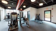 This single-floor fitness center is thoughtfully designed to support a comprehensive range of workout routines. The space is divided into distinct functional zones, each outfitted with professional-grade equipment to meet varied training needs.

The main exercise area features an exposed black ceiling with open ductwork — including a bold red ventilation duct — that gives the space an industrial character, complemented by large windows that flood the floor with natural light and offer views of the surrounding neighborhood. Pendant-style drum lights provide additional overhead illumination throughout.

The cardio zone is lined with treadmills, elliptical trainers, and stationary bikes positioned facing the windows, creating an energizing environment for endurance training. Adjacent to this, a strength training area houses Precor selectorized machines, a multi-function cable station, and an adjustable weight bench paired with a full dumbbell rack.

A dedicated functional training room offers a more enclosed setting with a drop ceiling, recessed lighting, and tiled walls. This area is equipped with an Octagon power rack, TRX suspension trainers, kettlebells, medicine balls, a barbell, and a plyo box — ideal for dynamic and high-intensity workouts. A rowing machine adds to the versatility of this zone.

Rubber flooring throughout ensures safety and durability, while a water fountain and restroom access add convenience for users.