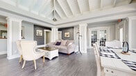 Located at 1631 Hill Rd N in Pickerington, Ohio, this single-floor home blends farmhouse charm with modern updates throughout its open, airy layout. The living area is a true centerpiece, showcasing a dramatic vaulted ceiling with exposed white-painted wood beams and tongue-and-groove planking that draws the eye upward and creates a sense of expansive height. Gray-toned hardwood-style flooring runs throughout, complementing the cool gray painted brick accent walls and crisp white architectural columns that define the space. Large windows on multiple walls flood the interior with natural light, offering views of the surrounding greenery. The dining area is bathed in sunlight through a generous bank of double-hung windows, providing a bright and inviting atmosphere for gatherings. A set of French doors connects the living space to an adjacent area, enhancing the open flow of the floor plan. The kitchen is outfitted with white shaker-style cabinetry, matte black hardware, white quartz countertops, and stainless steel appliances including a gas range, dishwasher, microwave, and refrigerator. A pass-through window connects the kitchen to the main living space, reinforcing the home's open and connected feel. Together, these spaces create a cohesive, light-filled interior with a warm yet refined farmhouse aesthetic.