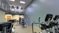 Located at 7303 Hihenge Ct in Raleigh, NC, this two-story fitness facility offers a well-appointed workout environment designed to accommodate a variety of exercise needs. The main floor features an open layout with a generous selection of strength training equipment, including cable machines, weight benches, and resistance stations, as well as cardio equipment such as elliptical trainers and cycling machines. A centrally positioned staircase with metal railings connects the ground level to the upper mezzanine, which is outfitted with additional strength machines visible from below. The open mezzanine design creates a sense of spaciousness and visual continuity between floors, enhanced by high ceilings that allow natural light to filter through recessed ceiling panels. The neutral color palette — featuring soft gray walls and light-toned flooring — contributes to a clean, functional atmosphere throughout the space. The layout is thoughtfully organized to maximize usable floor area while maintaining clear pathways between equipment. Overall, the facility presents a professional and well-maintained environment suited for a full range of fitness activities.