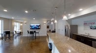 This single-floor space is designed with an open-concept layout that integrates the kitchen, dining, and living areas into one cohesive environment. The kitchen features a substantial granite-topped island with a white base, stainless steel refrigerator, and a trio of stylish pendant lights that hang overhead, providing both task lighting and a decorative focal point. The polished concrete floors extend throughout the entire space, lending an industrial yet refined character to the interior.

The dining area accommodates a compact table with sleek bar-height chairs and is complemented by a wall-mounted flat-screen television, making it a functional space for both meals and entertainment. Recessed lighting is distributed evenly across the ceiling, while wall sconces add warm accent lighting throughout. The white walls and neutral tones create a clean, bright atmosphere that enhances the open feel of the layout.

Further into the space, the open floor plan reveals additional areas, suggesting generous room for movement and versatile use. The overall design balances modern industrial elements with everyday functionality, resulting in a practical yet visually cohesive living environment.