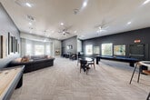 Located at 6800 Gaston Road in Katy, TX, this single-floor amenity space offers a well-appointed community lounge and game room designed for both relaxation and recreation. The open layout is unified by a distinctive herringbone-patterned carpet that runs throughout, complementing the contemporary color palette of cool grays, deep charcoal, and warm accent tones.

The lounge area features a tufted sectional sofa accented with warm-toned throw pillows, framed mirrors, and decorative wall art, all bathed in natural light from large windows with white shutters. An elegant pendant chandelier anchors the seating nook, adding a refined touch to the space.

The game area is a standout highlight, equipped with a full-size pool table, a shuffleboard table, and a round bistro-style table with seating — ideal for social gatherings. A wall-mounted flat-screen TV adds entertainment value to this zone.

A dedicated workstation area lines one wall, outfitted with computer monitors and counter-height seating, providing a functional workspace within the communal setting. Recessed lighting and a ceiling fan ensure the space remains comfortable throughout. Large windows along multiple walls allow natural light to flow in while offering views of the surrounding landscape, contributing to the bright and inviting ambiance of this versatile community space.