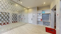 This building lobby serves as an elegant and functional point of entry, finished throughout with large-format marble-effect porcelain floor tiles that lend a polished, upscale aesthetic to the space. The lobby is thoughtfully organized into distinct functional zones. Near the main entrance, floor-to-ceiling glass doors provide views of the surrounding streetscape, allowing natural light to filter in and create a bright, welcoming atmosphere. A striking decorative glass partition wall featuring a geometric lattice pattern serves as a bold architectural focal point, adding visual interest and a contemporary design sensibility to the interior. Adjacent to this feature wall, a stainless steel elevator with marble-clad surrounds offers vertical access to the building's upper floors. On the opposite side of the lobby, a well-organized mailroom station includes a built-in shelving unit housing individual tenant mailboxes, open cubbies for package storage, and dedicated postal service areas. A stairwell access door, clearly signed, is conveniently located nearby. Recessed ceiling lighting throughout ensures the space is evenly and effectively illuminated. The overall ambiance is clean, modern, and professionally appointed, reflecting a well-maintained residential building with thoughtful attention to both form and function.