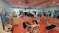 This fitness center is a thoughtfully designed, single-level facility offering a comprehensive range of equipment for both strength training and cardiovascular exercise. The space is defined by its vibrant red rubberized flooring, recessed lighting, and multiple ceiling fans that contribute to a comfortable workout environment.

The strength training area is generously appointed with a wide variety of white-framed machines, free-weight stations, adjustable benches, and multiple barbells loaded with weight plates. A tiered dumbbell rack provides an organized selection of free weights, while several cable and resistance machines accommodate a full range of muscle groups. Full-length mirrors line one wall, visually expanding the space and allowing users to monitor their form throughout their workout.

The cardio section houses a row of treadmills, stationary bikes, ellipticals, and stair climbers, positioned near windows that allow natural light to filter in. Wall-mounted televisions are positioned throughout the facility for entertainment during workouts.

A dramatic floor-to-ceiling panoramic mural of the Chicago skyline serves as a striking focal point, adding visual energy and a sense of place to the space. The overall ambiance is clean, functional, and energetic, making it a well-rounded destination for a variety of fitness goals.