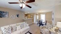 Located at 505 Regency Dr in Fayetteville, North Carolina, this single-floor residence offers a cohesive and functional layout designed for comfortable everyday living. The open-concept main area seamlessly connects the living room, dining area, and kitchen, creating a spacious and airy feel throughout. The living room is anchored by plush carpeting and features a ceiling fan with a light fixture overhead, providing both comfort and ambient lighting. Neutral-toned walls complement the warm, inviting atmosphere of the space. A breakfast bar-style counter serves as a natural divider between the living area and the kitchen, which is visible in the background and includes standard appliances. The dining area is bathed in natural light from a large window dressed with light curtains, and is accented by a chandelier overhead, adding a touch of elegance. White wainscoting detail along the dining area walls adds a classic architectural touch. The space benefits from recessed lighting and additional overhead fixtures throughout, ensuring well-lit interiors. The textured ceiling and consistent flooring tie the rooms together cohesively, making this a well-appointed, move-in ready single-floor home.
