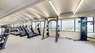 Located at 1254 Hennepin Ave N in Minneapolis, Minnesota, this fitness center offers a generously sized, single-floor workout facility designed to accommodate a wide range of exercise needs. The open floor plan is thoughtfully organized into distinct functional zones, creating an efficient and comfortable environment for users of all fitness levels.

The cardio section is extensively equipped, featuring a long row of treadmills, StairMaster steppers, elliptical machines, and stationary cycling bikes, each outfitted with individual display screens. A dedicated strength training area houses adjustable weight benches, a multi-station cable machine, a power rack, and a full dumbbell rack alongside medicine balls and stability equipment. Built-in open shelving with warm wood tones and blue accent walls near the entrance adds a welcoming, organized touch, complemented by a conveniently placed water station.

Throughout the space, large windows line the perimeter walls, flooding the interior with natural light and offering expansive views of the Minneapolis skyline and surrounding neighborhood. The clean, neutral palette — featuring light carpet flooring, white walls, and linear recessed and strip lighting along the ceiling — creates a bright, modern aesthetic. The well-planned layout ensures ample open floor space for movement between equipment zones, making this fitness center both functional and visually inviting.