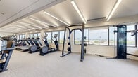 Located at 1254 Hennepin Ave N in Minneapolis, Minnesota, this fitness center offers a generously sized, single-floor workout facility designed to accommodate a wide range of exercise needs. The open floor plan is thoughtfully organized into distinct functional zones, creating an efficient and comfortable environment for users of all fitness levels.

The cardio section is extensively equipped, featuring a long row of treadmills, StairMaster steppers, elliptical machines, and stationary cycling bikes, each outfitted with individual display screens. A dedicated strength training area houses adjustable weight benches, a multi-station cable machine, a power rack, and a full dumbbell rack alongside medicine balls and stability equipment. Built-in open shelving with warm wood tones and blue accent walls near the entrance adds a welcoming, organized touch, complemented by a conveniently placed water station.

Throughout the space, large windows line the perimeter walls, flooding the interior with natural light and offering expansive views of the Minneapolis skyline and surrounding neighborhood. The clean, neutral palette — featuring light carpet flooring, white walls, and linear recessed and strip lighting along the ceiling — creates a bright, modern aesthetic. The well-planned layout ensures ample open floor space for movement between equipment zones, making this fitness center both functional and visually inviting.
