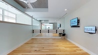This single-floor studio space is thoughtfully designed to support movement and wellness activities. The expansive open floor plan is finished with light-toned hardwood flooring that lends warmth and a clean aesthetic throughout the space. Full-length mirrors line the walls, visually expanding the room and serving as a practical feature for participants to monitor their form and technique. Mounted ballet barres extend along the mirrored wall, making this studio well-suited for dance, barre, yoga, or fitness classes. A wall-mounted flat-screen display provides a modern touch, enabling instructors to share visual content or stream programming for class participants. Natural light filters into the space through large windows that offer views to the exterior greenery, creating a refreshing and energizing atmosphere. The ceiling features a recessed acoustic tile panel alongside a ceiling fan, contributing to both sound management and air circulation. Recessed lighting and security cameras are also present, ensuring a well-lit and secure environment. Glass partition panels along one side of the studio add transparency and a contemporary architectural element. The overall ambiance is clean, airy, and professionally appointed, making it an inviting and functional setting for group fitness or wellness activities.