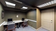 This single-floor office space is designed with functionality and practicality in mind. The interior features olive-green and taupe walls accented by white baseboards, creating a subdued, professional atmosphere. The dark ceiling panel contrasts with the recessed and drop-in fluorescent lighting fixtures that illuminate the space efficiently, compensating for the absence of natural light sources.

The main area is equipped with a built-in or freestanding light-colored workstation desk that runs along one wall, accommodating multiple computer workstations complete with desktop towers, monitors, keyboards, and task chairs with wooden frames. This setup suggests the space is configured for shared or multi-user computer access, suitable for administrative, clerical, or public-use purposes.

A framed certificate or document is displayed on the wall near the workstation, lending a degree of professionalism to the setting. A door on the right side of the room provides access to an adjoining space or exit. The patterned low-pile carpet adds a degree of texture underfoot while remaining durable and office-appropriate. Overall, the space is compact, well-organized, and oriented toward focused, task-driven work.