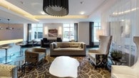 This community lounge is a thoughtfully designed gathering space that blends luxury finishes with functional amenities across a single, open floor plan. The lounge is organized into several distinct zones that flow seamlessly into one another, creating a versatile environment suited for both relaxation and socializing.

The main seating area is anchored by plush sofas, wingback chairs, and patterned accent chairs arranged atop an elegant geometric area rug. A sculptural white coffee table serves as a striking centerpiece, while a statement chandelier with a dramatic cylindrical silhouette draws the eye upward. Large windows dressed with floor-length drapes invite abundant natural light and outdoor views.

Adjacent to the seating area, a warming fireplace paired with a wall-mounted television creates an inviting focal point. A decorative lattice-style partition with a honeycomb pattern elegantly separates the lounge from a fully appointed kitchen, which features dark cabinetry, rich stone countertops, pendant lighting, and a herringbone-patterned floor.

A dedicated workstation area along the windowed wall offers desks and seating for residents needing a productive space. Near the entrance, a dramatic floor-to-ceiling textured glass water feature wall adds a tranquil, architectural element, complemented by herringbone hardwood flooring and warm ambient lighting throughout.