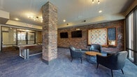 This well-appointed game room offers a dynamic blend of industrial design and modern comfort, making it an ideal space for entertainment and social gatherings. Exposed brick walls and structural brick columns add a distinctive character to the room, contrasting beautifully with the smooth, painted ceilings and blue-toned patterned carpet that runs throughout.

The space is divided into several functional zones. A billiards area takes center stage, anchored by a full-size pool table with cue sticks mounted on the adjacent wall. Nearby, an arcade cabinet flanked by upholstered stools and a wall-mounted flat-screen TV provides additional gaming options. A dedicated lounge zone features sleek leather club chairs arranged around a round coffee table, accompanied by two flat-screen TVs mounted on the exposed brick wall and a large decorative Scrabble board, adding a playful touch.

On the opposite end, floor-to-ceiling glass doors and oversized windows flood the space with natural light and open to an outdoor patio area with views of palm trees and surrounding architecture. This seating area includes additional leather chairs, a sputnik-style chandelier overhead, and wall-mounted cue storage. Bold abstract artwork adorns the walls near the pool table, completing the room's vibrant, urban aesthetic.