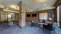 This well-appointed game room offers a dynamic blend of industrial design and modern comfort, making it an ideal space for entertainment and social gatherings. Exposed brick walls and structural brick columns add a distinctive character to the room, contrasting beautifully with the smooth, painted ceilings and blue-toned patterned carpet that runs throughout.

The space is divided into several functional zones. A billiards area takes center stage, anchored by a full-size pool table with cue sticks mounted on the adjacent wall. Nearby, an arcade cabinet flanked by upholstered stools and a wall-mounted flat-screen TV provides additional gaming options. A dedicated lounge zone features sleek leather club chairs arranged around a round coffee table, accompanied by two flat-screen TVs mounted on the exposed brick wall and a large decorative Scrabble board, adding a playful touch.

On the opposite end, floor-to-ceiling glass doors and oversized windows flood the space with natural light and open to an outdoor patio area with views of palm trees and surrounding architecture. This seating area includes additional leather chairs, a sputnik-style chandelier overhead, and wall-mounted cue storage. Bold abstract artwork adorns the walls near the pool table, completing the room's vibrant, urban aesthetic.