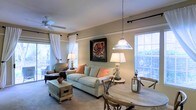 This single-story residence offers a thoughtfully arranged floor plan that makes efficient use of its space across several well-defined rooms. The layout, as seen from the dollhouse view, includes multiple bedrooms, living areas, a kitchen, and bathrooms arranged in an L-shaped configuration that promotes a natural flow throughout the home.

The living area serves as the heart of the space, featuring large windows dressed with white sheer curtains and horizontal blinds that allow generous natural light to fill the room. A ceiling fan overhead ensures comfort, while a pendant light anchors the dining area nearby. Warm, neutral wall tones complement the soft carpeted flooring, creating a cozy and welcoming atmosphere. The living room accommodates a comfortable seating arrangement centered around a light upholstered sofa accented with coral-toned throw pillows, alongside a rustic coffee chest that adds character.

The kitchen, visible in the floor plan, features white cabinetry and appliances in a compact yet functional configuration. Additional rooms include multiple bedrooms and bathrooms, each maintaining the home's consistent warm palette. Overall, the space presents a cohesive, comfortable interior with a relaxed, neutral aesthetic throughout.