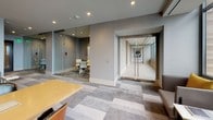 This sophisticated single-floor office suite is designed with a refined, contemporary aesthetic that balances professionalism with comfort. The space is anchored by a central open area featuring a large conference table surrounded by leather chairs, making it ideal for team collaboration and client meetings. Geometric, multi-tone carpeting in soft grays unifies the layout and adds visual depth throughout.

Glass-walled private offices and meeting rooms line the perimeter, providing transparency while maintaining acoustic separation. Textured neutral wallcovering and recessed lighting contribute to a polished, upscale atmosphere. A corridor with frameless glass doors connects zones seamlessly, enhancing the sense of openness and flow.

A standout feature of the space is the lounge area, which is bathed in natural light through expansive floor-to-ceiling windows. Comfortable gray armchairs accented with bold yellow cushions are positioned to take full advantage of sweeping views of an adjacent outdoor terrace. The terrace itself is outfitted with lounge chairs, planters, and string lighting, extending the usable space beyond the interior.

Gold-toned accent furniture, wall-mounted displays, and carefully curated furnishings throughout reflect a high level of finish and attention to detail. This space is well-suited for a professional office environment that prioritizes both function and elevated design.