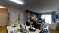 This single-floor residence offers a thoughtfully designed open-concept layout that integrates the kitchen, dining, and living areas into one cohesive, functional space. The kitchen is appointed with granite-look countertops, a double-basin stainless steel sink, and a flush-mounted ceiling light that brightens the workspace. A breakfast bar counter creates a natural transition into the dining area, which features a dark wood table paired with white upholstered chairs for a striking contrast. The living area beyond is anchored by a bold dark accent wall that adds depth and dimension to the space, complemented by neutral tones and patterned area rugs. A large window dressed with vertical blinds allows generous natural light to fill the room, creating a bright and welcoming atmosphere. Decorative touches, including fresh greenery and curated accessories, lend the space a warm, inviting feel. A doorway off the main living area leads to what appears to be a utility or laundry room, adding practical functionality. The overall design balances contemporary style with everyday comfort, making it a versatile and well-appointed living environment.