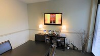 This single-floor professional space is thoughtfully arranged to provide a welcoming and informative atmosphere. The room features neutral, light-toned walls accented by a classic chair rail molding, lending a clean and polished aesthetic throughout.

A large flat-screen television is prominently wall-mounted, currently displaying content referencing Downtown D.C. and Rockville, suggesting a local real estate or hospitality-related use. Below the screen, a dark wood console unit holds an array of brochures, informational materials, and decorative items, flanked by two matching table lamps that cast a warm, inviting glow.

The flooring consists of a patterned low-pile carpet, adding texture while maintaining a professional appearance. A dark leather seating arrangement is partially visible in the foreground, indicating a comfortable area for guests or clients. Potted plants placed near the console introduce a subtle natural element, softening the overall look of the space.

The layout is efficient and organized, making it well-suited as a reception area, waiting room, or client-facing office. The combination of functional display areas, ambient lighting, and carefully chosen furnishings creates a space that is both informative and inviting for visitors.
