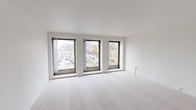 This well-appointed single-floor urban unit offers a clean, modern aesthetic throughout its thoughtfully designed layout. The spacious living area is a standout feature, bathed in abundant natural light from a trio of large windows framing elevated views of the surrounding cityscape, including a neighboring historic church and active street below. Neutral-toned carpet flows throughout the living and bedroom spaces, complementing the crisp white walls and smooth ceilings.

The bedrooms are generously sized, each featuring ceiling fans and large windows that allow natural light to filter in while offering views of the urban surroundings. Baseboard heating runs along the walls, ensuring year-round comfort.

The kitchen is efficiently designed with rich dark cabinetry, light stone-finish countertops, and stainless steel appliances including a refrigerator, electric range, and built-in microwave. A tall narrow window brings in a touch of natural light, adding to the kitchen's functional appeal.

The unit includes two bathrooms: a full bath featuring a tub-shower combination, a dark wood vanity with a white basin, classic mosaic tile flooring, and a mirror with overhead lighting; and a half bath with a pedestal sink and towel bar. The overall ambiance is clean, contemporary, and move-in ready.