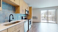 This thoughtfully designed single-floor unit offers a clean, contemporary aesthetic throughout. The kitchen is a standout feature, showcasing light wood cabinetry with matte black hardware, a striking blue hexagonal tile backsplash, and white quartz countertops. A full suite of stainless steel appliances — including a refrigerator, electric range, over-the-range microwave, and dishwasher — provides everything needed for everyday cooking and entertaining. Ample upper and lower cabinetry offers generous storage space.

The open living area flows seamlessly from the kitchen, bathed in natural light from a large sliding glass door that leads to a private balcony with views of the surrounding neighborhood. Light-toned luxury vinyl plank flooring runs throughout, complementing the neutral white walls and creating a bright, airy atmosphere.

The bathroom maintains the same modern sensibility, featuring a light wood vanity with a white undermount sink, quartz countertop, a large mirror, and a glass-enclosed shower. Clean lines and neutral finishes give the space a polished, functional feel. Overall, this unit combines modern finishes, practical design, and natural light to create a comfortable and inviting living environment.