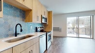 This thoughtfully designed single-floor unit offers a clean, contemporary aesthetic throughout. The kitchen is a standout feature, showcasing light wood cabinetry with matte black hardware, a striking blue hexagonal tile backsplash, and white quartz countertops. A full suite of stainless steel appliances — including a refrigerator, electric range, over-the-range microwave, and dishwasher — provides everything needed for everyday cooking and entertaining. Ample upper and lower cabinetry offers generous storage space.

The open living area flows seamlessly from the kitchen, bathed in natural light from a large sliding glass door that leads to a private balcony with views of the surrounding neighborhood. Light-toned luxury vinyl plank flooring runs throughout, complementing the neutral white walls and creating a bright, airy atmosphere.

The bathroom maintains the same modern sensibility, featuring a light wood vanity with a white undermount sink, quartz countertop, a large mirror, and a glass-enclosed shower. Clean lines and neutral finishes give the space a polished, functional feel. Overall, this unit combines modern finishes, practical design, and natural light to create a comfortable and inviting living environment.