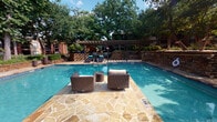 This outdoor amenity space centers around a generously sized swimming pool with clear blue water, framed by a natural stone deck laid in an irregular flagstone pattern. Wicker lounge chairs and side tables are positioned poolside, offering comfortable spots to relax and take in the surroundings. A robust dark wood pergola spans a significant portion of the deck, providing shaded areas beneath that include additional lounge seating and chaise lounges with teal cushions. Adjacent to the pergola is a well-appointed outdoor kitchen, featuring a built-in stainless steel grill set into a natural stone counter, along with a dining table and seating — ideal for al fresco gatherings. Layered stone retaining walls with warm, earthy tones define the perimeter of the pool area, adding a structured yet organic feel to the landscape. Vibrant cobalt blue ceramic planters punctuate the stonework with bold color accents. Mature oak trees with broad canopies arch overhead throughout the space, casting dappled natural light and creating a shaded, park-like atmosphere. The surrounding brick residential buildings are visible in the background, integrating the amenity seamlessly into the broader community environment.