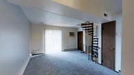 This two-floor space in Gray presents a practical and well-appointed layout designed for comfortable living. The main level greets visitors with a generously sized living area finished with soft blue-gray wall-to-wall carpeting that adds warmth underfoot. A large sliding glass door dressed with vertical blinds allows abundant natural light to fill the room while providing access to the outdoors. A wall-mounted air conditioning unit ensures year-round climate comfort, complemented by baseboard heating along the perimeter walls. Neutral white walls and a textured ceiling contribute to a clean, understated aesthetic throughout. A standout architectural feature of this space is the sleek black metal spiral staircase, which serves as both a functional connector to the upper floor and a visual focal point. Its compact, circular design makes efficient use of the floor plan while adding a contemporary industrial touch to the overall interior. The upper level is accessible via this eye-catching staircase, expanding the usable area of the home. Wood-toned doors provide classic contrast against the light walls. The overall ambiance is bright, open, and move-in ready, making this Gray property a versatile and inviting space.