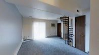 This two-floor space in Gray presents a practical and well-appointed layout designed for comfortable living. The main level greets visitors with a generously sized living area finished with soft blue-gray wall-to-wall carpeting that adds warmth underfoot. A large sliding glass door dressed with vertical blinds allows abundant natural light to fill the room while providing access to the outdoors. A wall-mounted air conditioning unit ensures year-round climate comfort, complemented by baseboard heating along the perimeter walls. Neutral white walls and a textured ceiling contribute to a clean, understated aesthetic throughout. A standout architectural feature of this space is the sleek black metal spiral staircase, which serves as both a functional connector to the upper floor and a visual focal point. Its compact, circular design makes efficient use of the floor plan while adding a contemporary industrial touch to the overall interior. The upper level is accessible via this eye-catching staircase, expanding the usable area of the home. Wood-toned doors provide classic contrast against the light walls. The overall ambiance is bright, open, and move-in ready, making this Gray property a versatile and inviting space.