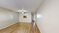 This single-floor residence offers a clean, functional layout with a cohesive neutral color palette and warm hardwood-style flooring that flows throughout the main living areas. The generous living room is brightened by natural light filtering through large windows dressed with blinds, and a ceiling fan adds both style and comfort overhead. A pass-through window connects the living area to the kitchen, enhancing the open feel of the space. The galley-style kitchen is efficiently designed with gray cabinetry lining both walls, laminate countertops, a stainless steel sink, an electric range, a dishwasher, and a tile mosaic backsplash accent. Light ceramic tile flooring completes the kitchen's practical finish. A hallway leads to the bedroom, which features its own ceiling fan, a sun-filled window, and dual reach-in closets with louvered bifold doors offering ample storage with built-in shelving and hanging rods. The bathroom includes a full-size soaking tub with an overhead shower, a vanity with cabinet storage, and tile flooring. Additional built-in shelving is visible in the hallway, providing convenient storage. Textured ceilings are present throughout, contributing to the space's classic residential character.