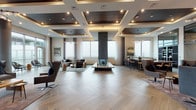 This expansive, single-floor lounge space showcases a refined contemporary design that seamlessly blends open-plan living with distinct functional zones. The signature herringbone-patterned hardwood flooring flows throughout, anchoring the entire space in warmth and visual continuity.

At the heart of the main gathering area stands a dramatic double-sided stone fireplace, clad in stacked slate tile, serving as a striking architectural focal point. Surrounding it, multiple seating arrangements offer versatile lounging options, from plush sectional sofas to tufted leather accent chairs. The coffered ceiling design, featuring dark wood insets and recessed lighting, adds depth and a layered sense of sophistication overhead.

A well-appointed bar and kitchen area features a large dark island with leather bar stools, complemented by a built-in wine display nearby. Integrated shelving units throughout the space provide decorative display and storage.

Floor-to-ceiling windows and glass walls flood the interior with natural light while framing sweeping city views. A dedicated seating nook with a stacked stone accent wall offers a quieter retreat. Glass doors open directly to an outdoor terrace with a pergola structure, extending the space's entertaining potential while maintaining an elevated urban atmosphere throughout.