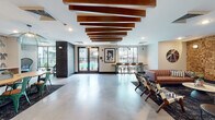 This expansive single-floor amenity space is thoughtfully designed to accommodate a variety of social and recreational activities. The open layout is unified by polished concrete flooring throughout and connects several distinct functional zones seamlessly.

The main lounge area features a striking ceiling installation of staggered wooden beams, adding warmth and architectural interest overhead. A seating area anchors this zone, centered around a textured mosaic wood-panel accent wall housing a wall-mounted television and a linear fireplace. Comfortable leather sofas and patterned accent chairs are arranged to encourage conversation and relaxation.

Adjacent to the lounge is a dining area distinguished by bold botanical wallpaper, a long communal table with vibrant green metal chairs, pendant lighting, and a kitchenette with cabinetry and a beverage refrigerator.

The game room is a dedicated entertainment hub equipped with a pool table beneath a statement pendant light, classic arcade machines, a foosball table, and built-in bench seating flanking a secondary television on another mosaic wood-panel wall. Large windows throughout the space invite natural light and offer views of a nearby outdoor pool area.