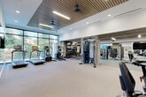 This contemporary fitness center is designed to deliver a comprehensive workout experience across a spacious, single-level layout. The main cardio area is anchored by floor-to-ceiling windows that flood the space with natural light and offer views of surrounding greenery, creating an energizing atmosphere. Treadmills, ellipticals, and a stair climber are arranged along this window wall, complemented by a distinctive slatted wood ceiling with integrated lighting and ceiling fans that add warmth to the modern design.

A functional training zone features a cable machine system, wall-mounted shelving stocked with medicine balls, resistance bands, and fitness mats, along with a heavy bag and large-screen TV. Full-length mirrors expand the sense of space while supporting form-conscious training. An adjoining mirrored room, enclosed by sleek black steel-framed glass partitions, houses cycling bikes and rowing machines for dedicated cardio sessions.

A separate strength training area with additional weight machines and dumbbells extends the facility's versatility. Near the entrance, deep-blue vertical tile accents the wall behind a dual water fountain and bottle-filling station. Built-in storage cabinetry and curated wall art complete the polished, upscale aesthetic throughout.