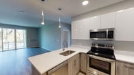 This single-floor unit showcases a contemporary design with a neutral color palette and thoughtful finishes throughout. The open-concept layout connects the kitchen and living area seamlessly, creating a bright and functional shared space. Light wood-tone vinyl plank flooring runs through the main living areas, adding warmth to the modern aesthetic.

The kitchen is well-appointed with a peninsula featuring quartz countertops, warm wood-toned lower cabinetry, and high-gloss white upper cabinets extending to the ceiling for ample storage. Stainless steel appliances — including a refrigerator, electric range, over-the-range microwave, and dishwasher — complement the space, while sleek pendant lights hang above the peninsula. The living area benefits from a sliding glass door that opens to a private balcony, allowing natural light and outdoor views to flow in.

The bedrooms are carpeted for comfort, each featuring large windows that bring in natural light, with at least one room offering a reach-in closet. Two bathrooms serve the unit: one with a soaking tub and subway tile surround, and another with a walk-in tiled shower, both featuring quartz vanity countertops and wood-toned cabinetry.