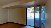 This single-floor space showcases a clean, modern design with smooth white walls and warm hardwood flooring that runs throughout the interior. A defining feature is the floor-to-ceiling sliding glass door, framed in sleek black metal, which serves as both a functional entryway and a dramatic architectural element. It floods the interior with natural light and provides direct access to a private outdoor patio enclosed by a white privacy fence. The patio extends the living area outdoors and overlooks surrounding greenery and neighboring buildings, giving the space an urban yet open feel. The interior maintains a minimalist aesthetic with neutral tones, creating a versatile canvas suitable for a variety of uses. A structural soffit runs along the ceiling near the sliding door, adding architectural interest while housing utilities discreetly. The baseboards and wall finishes are clean and well-maintained, contributing to the overall polished appearance. The combination of natural light, outdoor access, and contemporary finishes makes this a functional and inviting single-level space.