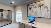 Located at 14723 W Oaks Plaza Dr in Houston, Texas, this single-story office suite delivers a refined and professional environment with thoughtfully curated design details throughout. The space is accessed through a striking black-framed glass door that opens into a corridor finished with herringbone-patterned hardwood flooring, setting an elegant tone from the moment of entry.

The primary workspace features built-in cabinetry with gray shaker-style doors topped with white marble-look countertops, providing ample workspace and storage. A large-scale abstract artwork anchors the wall above the workstation, adding a sophisticated artistic element to the room. Crown molding runs along the upper perimeter, lending a polished architectural detail to the space.

A standout architectural feature throughout is the arched window design — gracefully curved at the top and fitted with white wood blinds — which invites abundant natural light while offering views of the surrounding greenery. An additional office or reception area includes a built-in shelving unit with marble-top surfaces and an arched wall niche adorned with a decorative sunburst mirror, creating a welcoming and stylish focal point. Recessed lighting, neutral warm-gray walls, and plush carpet flooring complete the interior, resulting in a cohesive, upscale professional atmosphere.
