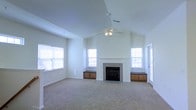This single-floor living space is defined by its vaulted ceiling, which lends an open, airy feel to the room. A ceiling fan is centrally mounted, providing both functionality and a finishing architectural detail that complements the angled roofline above. The room is finished with neutral wall-to-wall carpeting and crisp white walls, creating a clean and versatile backdrop.

The focal point of the space is a tiled fireplace centered along the back wall, framed by built-in wooden cabinetry with dark countertops on either side. These built-ins offer practical storage and display options while adding warmth and symmetry to the room's design.

Natural light flows generously through multiple windows positioned on both the front and sides of the room, including a door with glass paneling on the right side that may lead to an outdoor area. The window treatments are simple white blinds, keeping the aesthetic minimal and bright.

Overall, the space presents a welcoming, well-lit environment with thoughtful architectural details, ample storage, and a comfortable, functional layout suited for everyday living.