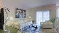 This well-appointed single-floor space offers a clean, contemporary aesthetic throughout. The living area serves as the centerpiece of the home, showcasing a light and airy atmosphere enhanced by neutral wall tones and plush carpeting. White upholstered seating, including a full sofa and accent chair, are artfully arranged around a distinctive glass-top coffee table with a sculptural dark wood base, lending a modern yet inviting feel to the room. A boldly patterned blue and white area rug anchors the seating arrangement, adding a vibrant touch of color and visual interest to the space. Two framed pieces of decorative artwork depicting elegant figures hang side by side on the main wall, contributing a cultured, refined ambiance. Natural light pours in through a large sliding glass door fitted with vertical blinds, offering both brightness and privacy. The door suggests access to an outdoor area, further enhancing the open, connected feel of the space. Recessed ceiling vents and smooth white ceilings complete the polished interior, while a soft table lamp adds warm accent lighting. Overall, the space presents a harmonious blend of comfort and style in a bright, well-designed layout.