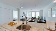 This single-floor residence is designed with an open-concept layout that seamlessly connects the kitchen, dining, and living areas into one cohesive, light-filled space. The kitchen is anchored by a generous island topped with elegant marble-style stone countertops featuring soft veining, paired with a sleek stainless steel undermount sink and a contemporary high-arc faucet. A glass pendant light hangs above the island, adding a refined decorative touch.

The open living and dining area beyond the kitchen is bathed in natural light, courtesy of large windows that frame views of lush greenery outside. The bright white walls and light-toned ceilings enhance the airy, expansive feel of the space. The living area is furnished with a neutral sofa and accented by modern décor, while a glass-topped dining table with dark chairs adds a sophisticated contrast.

Visible through an open doorway, the bedroom continues the clean, minimalist aesthetic with white bedding and soft, neutral tones. Throughout the residence, the consistent palette of whites and naturals creates a calm, contemporary ambiance that feels both welcoming and refined.