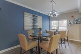 Located at 553 Settlers Landing in Hampton, Virginia, this single-floor community space is thoughtfully designed with a cohesive coastal-inspired color palette and modern furnishings. The layout flows seamlessly between distinct functional areas, all unified by warm hardwood-style flooring, crown molding, and bold teal accent walls that lend the space a sophisticated yet welcoming ambiance.

The lounge area serves as a central gathering point, featuring a comfortable gray sofa accented with navy and chartreuse throw pillows, flanked by tufted wingback chairs atop a striking blue-and-white patterned area rug. Abstract artwork adorns the deep teal feature wall, adding a dynamic artistic focal point. A quieter seating nook nearby offers two upholstered chairs beside a large window that invites ample natural light, creating an ideal spot for relaxed conversation or reading.

The dining area accommodates multiple guests with two high-top tables and upholstered counter-height chairs, lit by a modern globe chandelier. The shared teal accent wall continues here, maintaining visual continuity throughout the open layout.

The kitchen is well-appointed with warm wood cabinetry, granite countertops, tile flooring, and a full suite of stainless steel appliances, including a refrigerator, range, and built-in microwave — making it fully functional for events and gatherings.
