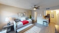 This single-floor studio unit offers a thoughtfully designed open layout that efficiently combines living, sleeping, and dining functions within one cohesive space. Light gray walls and wood-look plank flooring run throughout, creating a clean, contemporary aesthetic. A textured ceiling with a sleek ceiling fan provides both style and comfort to the main living area.

The primary living and sleeping area is generously proportioned, accommodating a daybed with trundle, a dresser, and a compact desk workspace positioned near a window that allows natural light to filter in. The window is fitted with horizontal blinds for privacy, while views of the surrounding exterior are visible through the glass.

Adjacent to the main room is a compact kitchenette featuring white cabinetry, light countertops, and bar-style seating, providing a practical space for meal preparation and casual dining.

The bathroom is well-finished with a marble-style vanity countertop, an inset sink, chrome fixtures, and a large mirror framed by overhead lighting — offering a clean and functional design. The bathroom opens directly to the main living area, reinforcing the studio's connected, open feel.

Overall, this studio presents a practical and modern living environment, making efficient use of its single-floor footprint with attention to both functionality and style.