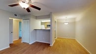 This single-story unit at Parkway Square offers a well-organized layout with distinct living, kitchen, bedroom, and bathroom areas. The living room is a comfortable, open space finished with wood-style laminate flooring and a ceiling fan with light fixture. A sliding glass door with vertical blinds allows natural light to fill the room and provides direct access to an outdoor area with greenery visible beyond. A pendant light fixture illuminates a secondary area adjacent to the living room, which connects seamlessly to the rest of the unit.

The kitchen is designed in a galley configuration, featuring light wood cabinetry, granite-style laminate countertops, a double stainless steel sink, and white appliances including a refrigerator, electric range, and an over-the-range microwave. A pass-through counter with a raised bar connects the kitchen to the living area. Wood-style flooring continues throughout the kitchen.

The bedrooms are carpeted and generously proportioned, each offering natural light through windows with blinds. One bedroom includes a walk-in closet, while the other provides ample space with a standard closet. The bathroom features a granite-style vanity countertop with wood cabinetry, a wall-mounted mirror, and a separate shower area with wood-style flooring.