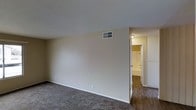 This single-floor residential unit offers a straightforward and functional layout designed for comfortable everyday living. The main living area is finished with wall-to-wall carpet, providing a soft underfoot feel and a warm, understated ambiance. Neutral-toned walls throughout the space create a clean, versatile backdrop suitable for a variety of furnishing styles.

Ceiling-mounted HVAC vents are integrated throughout, ensuring efficient climate control across the living areas. The textured ceiling adds a subtle architectural detail consistent with the overall practical design of the unit.

A doorway from the main living area connects to an adjacent hallway or secondary space, where the flooring transitions to dark hardwood-style planks, offering a visual contrast to the carpeted areas. This connecting corridor leads to what appears to be a bathroom, featuring light-colored walls and a bright, well-lit interior visible through the open doorway.

White trim and baseboards frame each room neatly, contributing to a cohesive and tidy aesthetic throughout. The unit's single-floor design ensures ease of navigation, making all areas conveniently accessible. Overall, the space presents a clean, move-in-ready environment with a no-frills, practical character suited to a wide range of occupants.