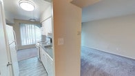 This single-floor residential unit offers a practical and well-organized layout designed for comfortable everyday living. The kitchen serves as a central feature of the space, outfitted with white cabinetry, a gas range, over-the-range microwave, refrigerator, and a double-basin sink with dark countertops. The kitchen is finished with light wood-style vinyl flooring, providing a clean and easy-to-maintain surface. A structural column partially separates the kitchen from the adjacent living area, maintaining an open feel while defining each zone. The living area features plush carpeting in a neutral tone, contributing to a warm and inviting atmosphere. Flush-mounted ceiling light fixtures provide consistent overhead illumination throughout. A window in the kitchen area allows natural light to flow in, brightening the space during daylight hours. Soft, neutral wall tones are carried throughout the unit, creating a cohesive and understated aesthetic. Additional hallway access visible from the kitchen connects to other areas of the unit, suggesting a thoughtfully arranged floor plan that balances openness with defined functional spaces.