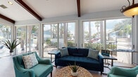 Located at 3099 Park Village Way in Melbourne, Florida, this single-story leasing office offers a welcoming and well-appointed environment designed to make a strong first impression. The space is characterized by soaring vaulted ceilings accented with rich, dark-stained exposed wood beams that add architectural warmth and character throughout.

Natural light floods the interior through expansive floor-to-ceiling sliding glass windows and doors that wrap around the main gathering area, providing unobstructed views of the surrounding palm trees, lush landscaping, and community pool. Light wood-tone flooring runs throughout, complementing the soft blue-gray wall palette and creating a cohesive, airy coastal aesthetic.

The lounge area is furnished with a deep navy sofa and teal accent chairs arranged around a woven rattan coffee table, offering a comfortable and inviting space for prospective residents. A ceiling fan with a gold finish and recessed lighting ensure the space remains bright and comfortable year-round.

A dedicated workstation area features a modern wood desk with ample workspace, accompanied by guest seating for one-on-one consultations. Wall-mounted artwork and decorative accents add personality to the professional setting. A flat-screen TV is mounted along one wall, and a charming arched entry door serves as a focal point at the main entrance, reinforcing the space's welcoming character.